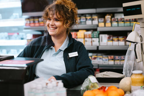 Glückliche Frau an der Kasse eines Supermarktes, die einen Kunden bedient. Frau, die an der Kasse eines Lebensmittelladens arbeitet, spricht mit einem Kunden und lächelt. - JLPSF11841