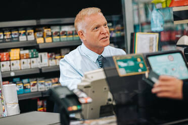 Älterer Mann, der an der Supermarktkasse arbeitet. Kassiererin im Gespräch mit einem Kunden, der mit Kreditkarte im Lebensmittelgeschäft bezahlt. - JLPSF11844