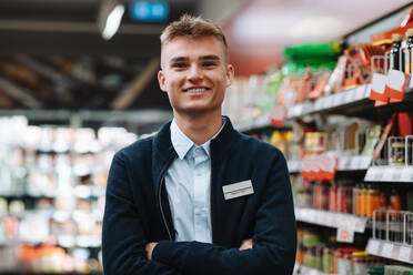 Porträt eines jungen Angestellten in einem Lebensmittelgeschäft, der lächelnd in die Kamera blickt. Ein Mann in Uniform steht mit verschränkten Armen im Supermarkt. - JLPSF11856