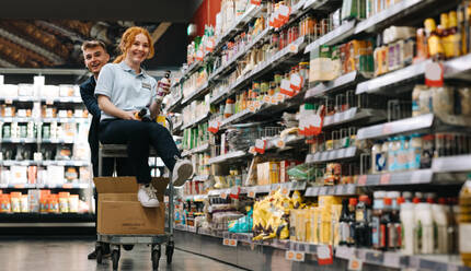 Zwei junge Arbeiter haben Spaß beim Einpacken von Waren. Eine Frau sitzt auf einem Einkaufswagen, der von einem männlichen Kollegen im Supermarktgang geschoben wird. - JLPSF11858