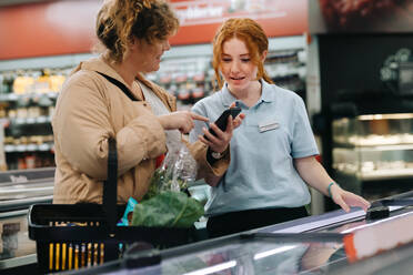 Mitarbeiter eines Lebensmittelgeschäfts hilft einer Kundin beim Einkaufen. Eine Kundin zeigt dem Verkäufer ihr Handy und fragt nach einem Produkt im Supermarkt. - JLPSF11860