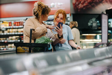 Eine Kundin zeigt dem Verkäufer ihr Handy und fragt nach den Produkten im Supermarkt. Eine Kundin im Lebensmittelgeschäft lässt sich von einer Mitarbeiterin helfen. - JLPSF11861