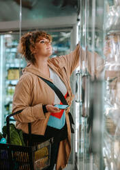 Female with shopping basket taking food from a fridge in grocery store. Woman customer buying groceries in supermarket. - JLPSF11872