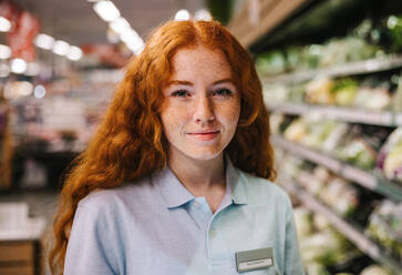Closeup of a young shop assistant. Female employee in uniform working in supermarket looking at camera. - JLPSF11877