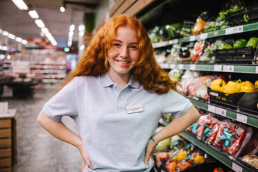 Portrait of a female worker at a grocery store and looking at the camera smiling. Woman in uniform standing with hands on hips in supermarket. - JLPSF11880