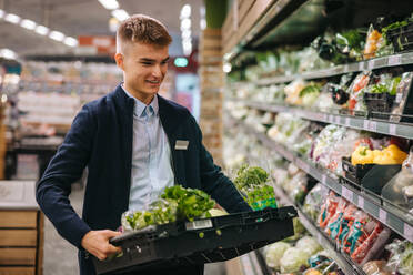 Verkäuferin im Supermarkt, die frisches Gemüse in die Regale der Gemüseabteilung einräumt. - JLPSF11883