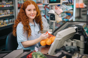 Woman cashier sitting behind checkout and working in supermarket. Female checkout counter clerk scanning grocery products and looking at camera. - JLPSF11891