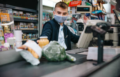 Young sales clerk wearing face mask sitting by cash register in supermarket and serving shoppers. Male cashier scanning grocery products at checkout. - JLPSF11898