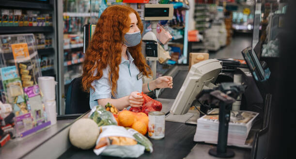 Kassiererin mit Gesichtsmaske in einem Supermarkt an der Kasse. Kassiererin beim Scannen von Lebensmitteln für Kunden während einer Pandemie. - JLPSF11899