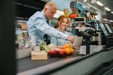 Senior manager helping new employee at checkout counter in supermarket. Mature man showing and explaining the checkout system to a trainee. - JLPSF11906