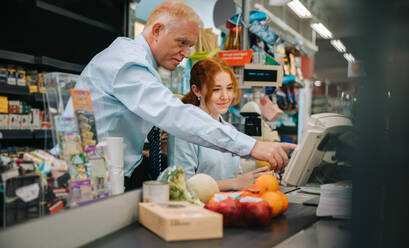 Senior manager training a new female employee at checkout counter. New cashier getting some help from store manager. - JLPSF11907