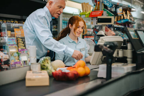 Supermarktleiter schult junge Mitarbeiterin an der Kasse. Älterer Mann unterrichtet neue Mitarbeiterin im Lebensmittelgeschäft über den Kassiervorgang. - JLPSF11908