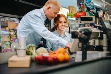 Senior manager giving training to a new female employee at checkout counter. Young worker getting some help from store manager at checkout counter. - JLPSF11909