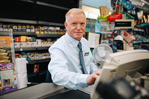 Ein älterer Mann sitzt an der Kasse eines Supermarktes und schaut in die Kamera, ein Supermarktleiter an der Kasse. - JLPSF11913