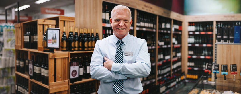 Porträt eines erfolgreichen Weinhändlers: Ein älterer Geschäftsmann steht mit verschränkten Armen in einem Weinladen und lächelt in die Kamera. - JLPSF11919