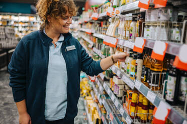 Eine Frau arbeitet im Supermarkt und ordnet die Produkte korrekt im Regal an und lächelt. - JLPSF11923