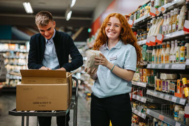 Junger Angestellter beim Auffüllen der Regale mit einem Kollegen im Supermarkt. Zwei Angestellte eines Lebensmittelgeschäfts arbeiten zusammen. - JLPSF11930