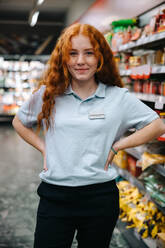 Kaukasische weibliche Angestellte bei der Arbeit im Supermarkt. Frau in Uniform steht in einem Lebensmittelladen mit ihren Händen auf den Hüften und schaut in die Kamera. - JLPSF11938