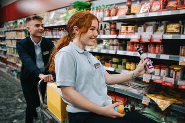 Zwei junge Arbeiter, die sich bei der Arbeit in einem Supermarkt amüsieren. Eine Frau sitzt auf einem Einkaufswagen, der Regale einräumt, und ein männlicher Kollege schiebt in einem Supermarkt-Gang. - JLPSF11945