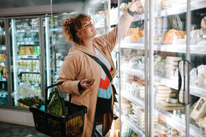 Frau beim Abholen von Produkten aus dem Regallager. Frau beim Einkaufen von Lebensmitteln im Supermarkt. - JLPSF11960