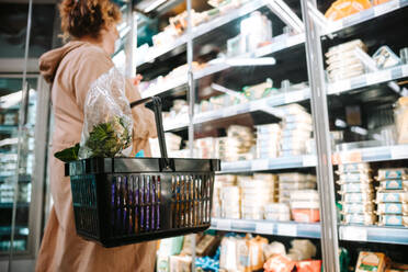 Woman holding a shopping basket and looking at grocery items in rack. Shopper buying groceries in supermarket. - JLPSF11962