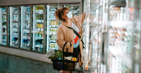 Frau beim Abholen von Produkten aus dem Regallager. Frau mit Gesichtsmaske beim Einkaufen im Supermarkt. - JLPSF11964