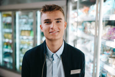 Junger Verkäufer in einem modernen Supermarkt. Ein Mann bei einem Ferienjob in einem Lebensmittelladen lächelt in die Kamera. - JLPSF11966