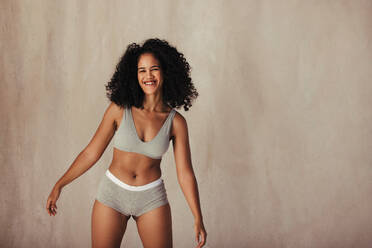 Pretty young female model enjoying being in her natural body. Body positive model wearing underwear and smiling at the camera while standing alone against a studio background. - JLPSF11974