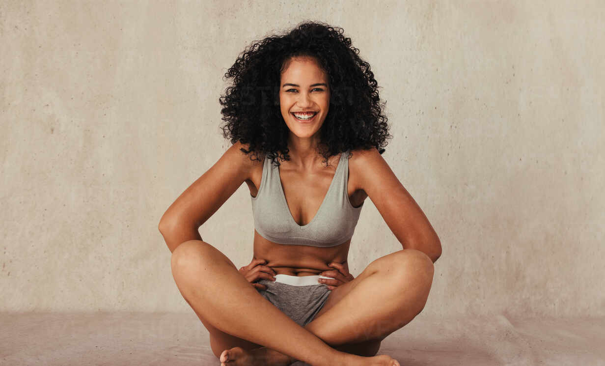 Having fun in underwear. Happy young woman smiling cheerfully while sitting  on the floor in a studio. Confident young woman embracing her natural body  while wearing beige underwear. stock photo