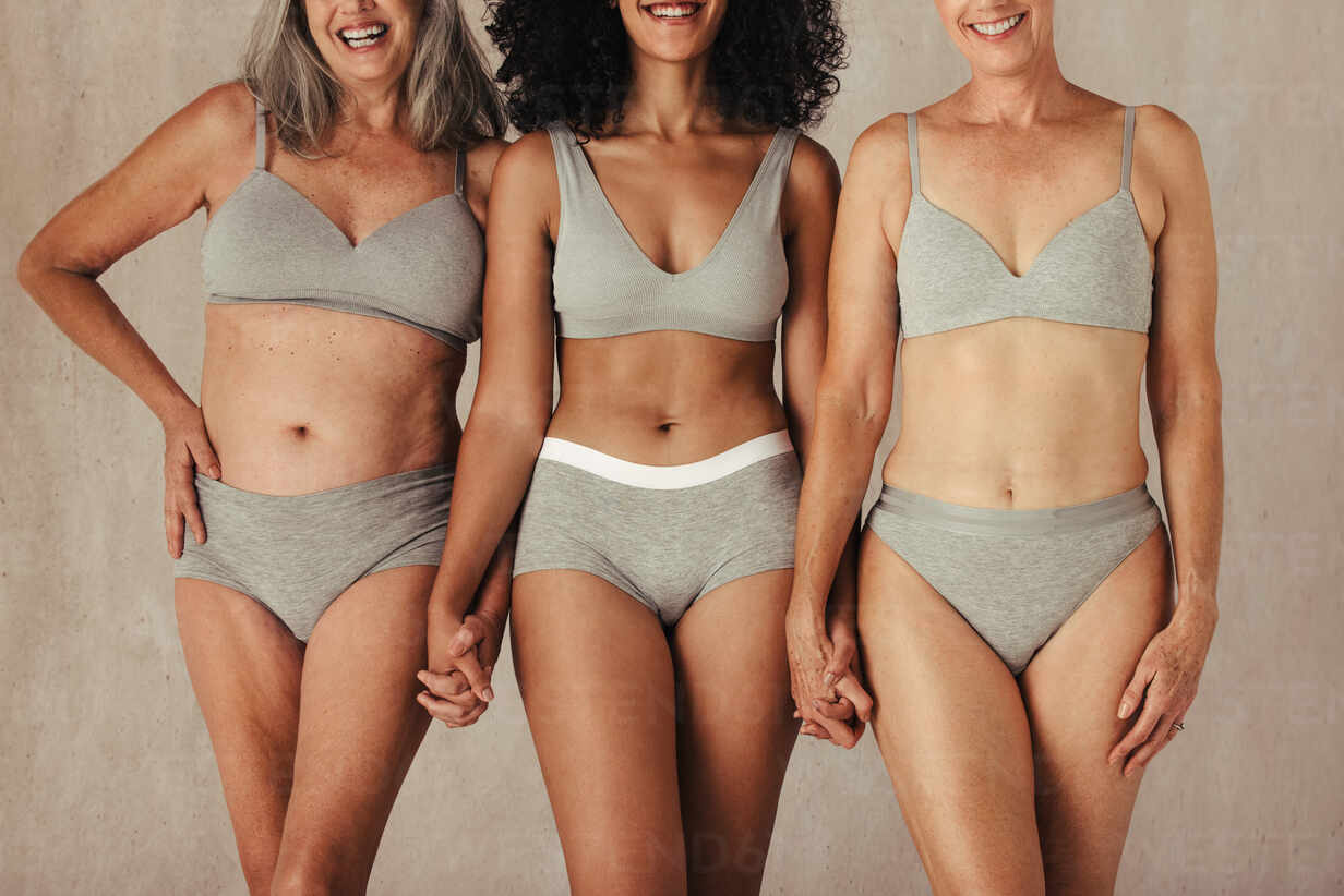 Unrecognizable natural female bodies in a studio. Diverse group of women in underwear  holding hands while standing together. Three women of different ages  smiling against a studio background. stock photo