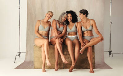 Glückliche Frauen unterschiedlichen Alters, die ihren natürlichen Körper in einem Studio umarmen. Vier körperbewusste Frauen, die Unterwäsche tragen und fröhlich lächeln. Frauen, die vor einem Studiohintergrund zusammensitzen. - JLPSF12002