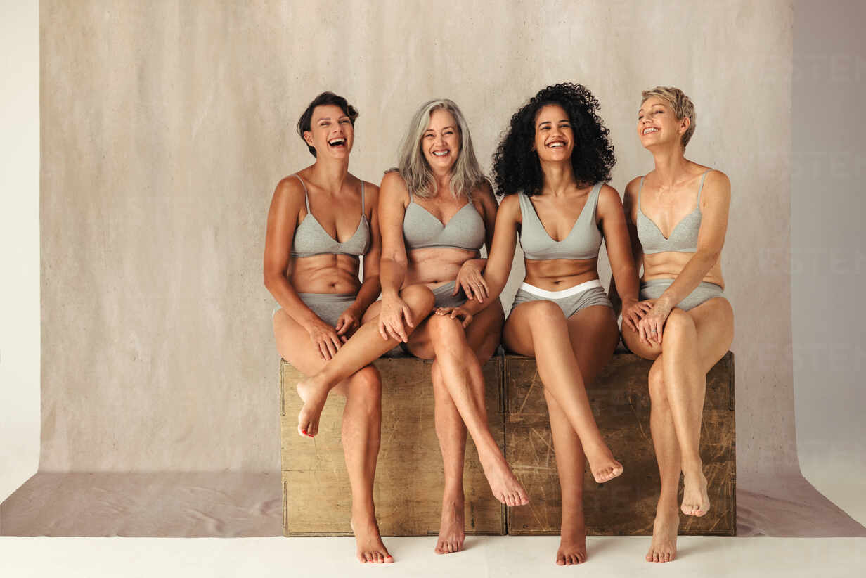Full body shot of four natural and body positive women of different ages  posing together. Confident women in underwear smiling and sitting together  against a studio background. stock photo