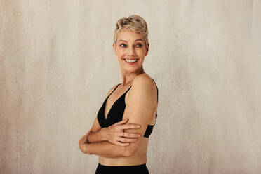 Carefree mature woman embracing her natural and aging body. Confident woman wearing black underwear and smiling cheerfully. Woman standing alone against a studio background. - JLPSF12030