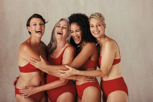 Four smiling women of different ages embracing their natural and aging bodies. Four happy and body positive women embracing each other while wearing red underwear against a studio background. - JLPSF12053