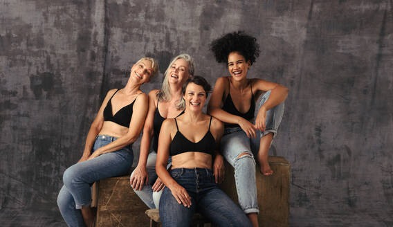 Vier körperbewusste Frauen unterschiedlichen Alters feiern ihre natürlichen Körper, während sie Jeans vor einem Studiohintergrund tragen. - JLPSF12063