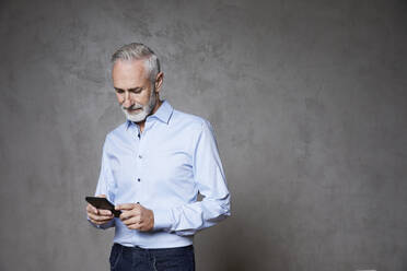 Älterer Mann mit Smartphone vor grauem Hintergrund - FMKF07739