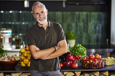 Happy man with arms crossed standing by kitchen counter - FMKF07767