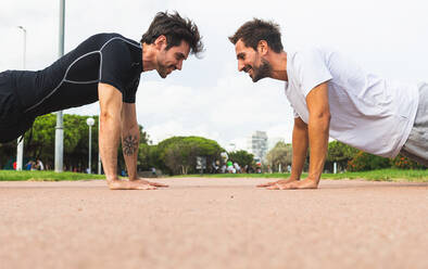 Seitenansicht eines hispanischen männlichen Sportlers, der sich bei einer Plank-Übung auf einem Pfad während eines Fitnesstrainings im Park ansieht - ADSF39819