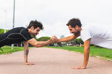 Seitenansicht von hispanischen männlichen Sportlern, die sich an den Händen fassen und sich gegenseitig ansehen, während sie eine Plank-Übung auf einem Pfad während eines Fitnesstrainings im Park machen - ADSF39820