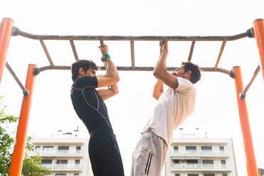 From below male athletes in sportswear doing pull up exercise on bars during fitness workout on sports ground in city - ADSF39821