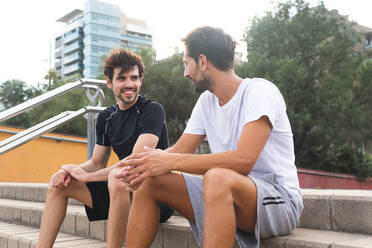 Niedriger Winkel von fröhlichen hispanischen Sportlern, die sich lächelnd ansehen, während sie auf Stufen sitzen und während einer Pause beim Fitnesstraining auf der Straße sprechen - ADSF39822