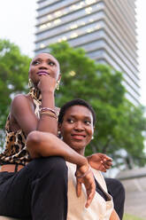 Niedriger Winkel von positiven erwachsenen stilvollen afroamerikanischen Freundinnen, die zusammen auf einer Stadtstraße sitzen und in der Nähe von modernen Gebäuden wegschauen - ADSF39836