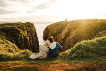 Back view of anonymous man and woman in outerwear leaning on each other and observing sea while sitting on cliff at sundown in Aviles, Spain - ADSF39837