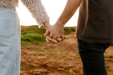 Anonymer Mann und Frau in Freizeitkleidung, die sich vor einem unscharfen Hintergrund mit Landschaft und Sonnenuntergang während eines romantischen Dates in Aviles, Spanien, an den Händen halten - ADSF39842