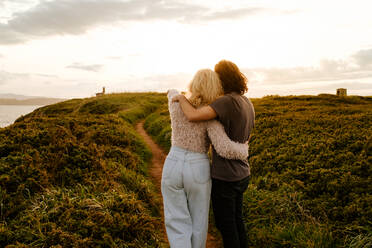 Rückenansicht blonde Frau und Mann, die sich umarmen und wegschauen, während sie auf einer grasbewachsenen Klippe bei Sonnenuntergang in Aviles, Spanien, stehen - ADSF39843