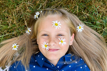 Top view of blond girl with fresh daisy flowers on face and hair looking at camera with smile while lying on grass on summer day - ADSF39846