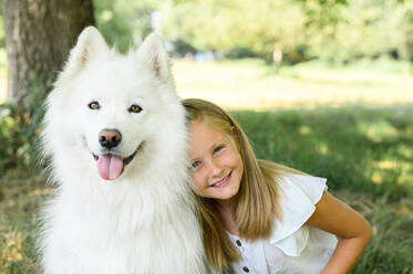 Fröhliches Mädchen, das ein gehorsames Samojeden-Mädchen umarmt und lächelnd in die Kamera schaut, während es im Sommer im Park Zeit verbringt - ADSF39847