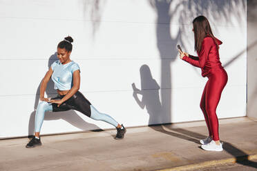 Full body of young athletic female in stylish sportswear taking photo of positive African American sportswoman doing lunges exercise against white wall on street - ADSF39857
