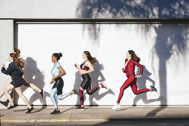 Seitenansicht ganzer Körper der jungen fit multiethnischen weiblichen Athleten in stilvollen Sportkleidung läuft auf dem Bürgersteig in der Nähe von weißen Wand während Outdoor-Fitness-Training am sonnigen Tag - ADSF39859