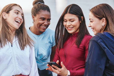 Group of happy sportswoman in activewear sharing news in social media with multiracial girlfriends during break in fitness workout on city street - ADSF39863